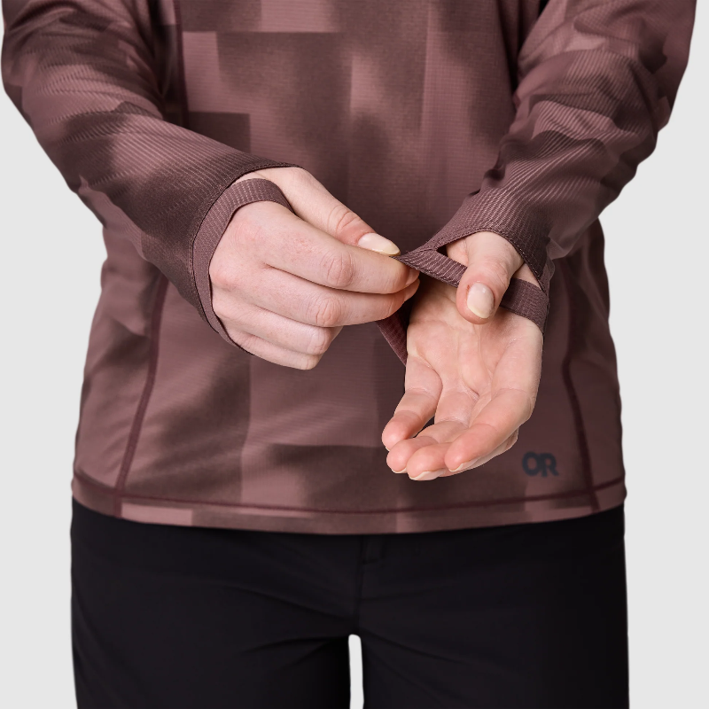 A close-up of a person wearing a brown athletic long-sleeve top and black pants, demonstrating how to fasten the adjustable wrist strap of the top.