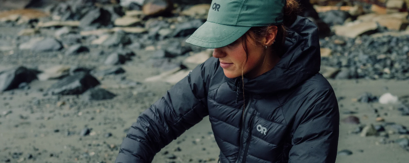 A woman wearing an orange outdoor puffer jacket and a matching green cap, sitting on a rocky beach.