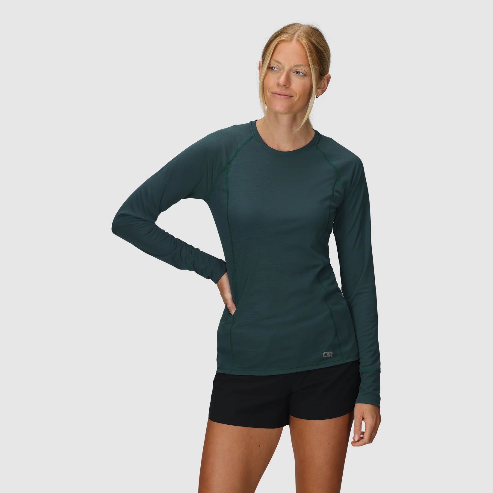 Woman wearing a green long-sleeve Outdoor Research hiking top and black hiking shorts on a white background