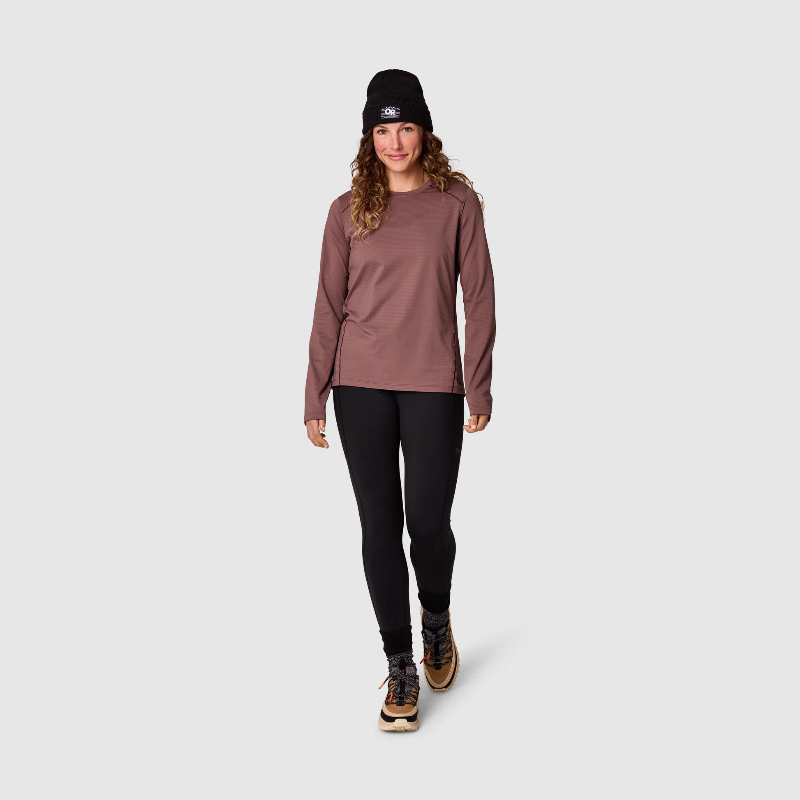 A woman modeling a long-sleeve athletic top in a dusty rose color, paired with black leggings, a black beanie, and hiking boots, against a light gray studio background.