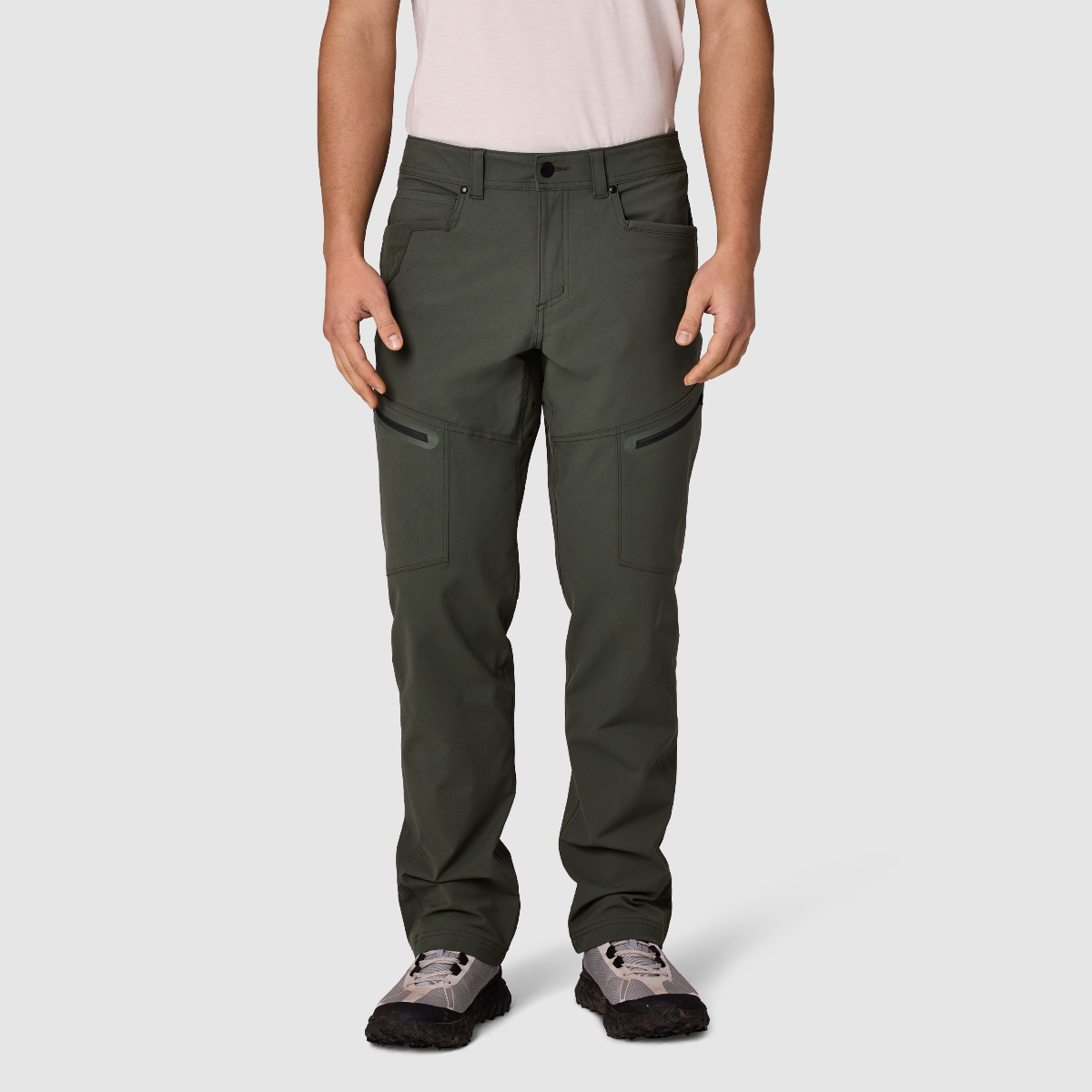 A person modeling a pair of dark olive green technical cargo pants with zippered side pockets, paired with a light pink t-shirt and gray hiking shoes, against a light gray studio background.