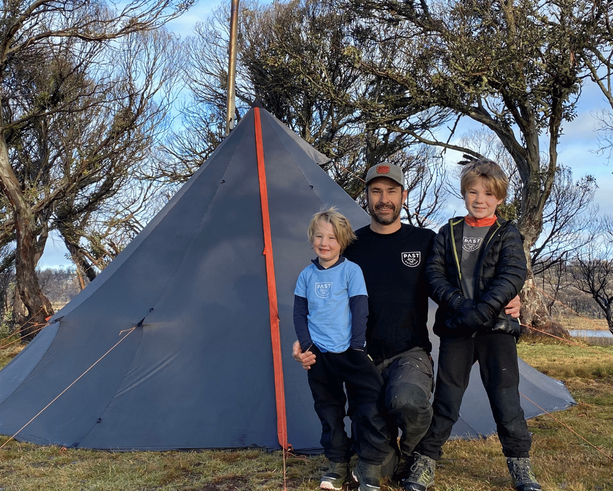 Three people standing in front of a tent in a natural setting with trees and blue sky.