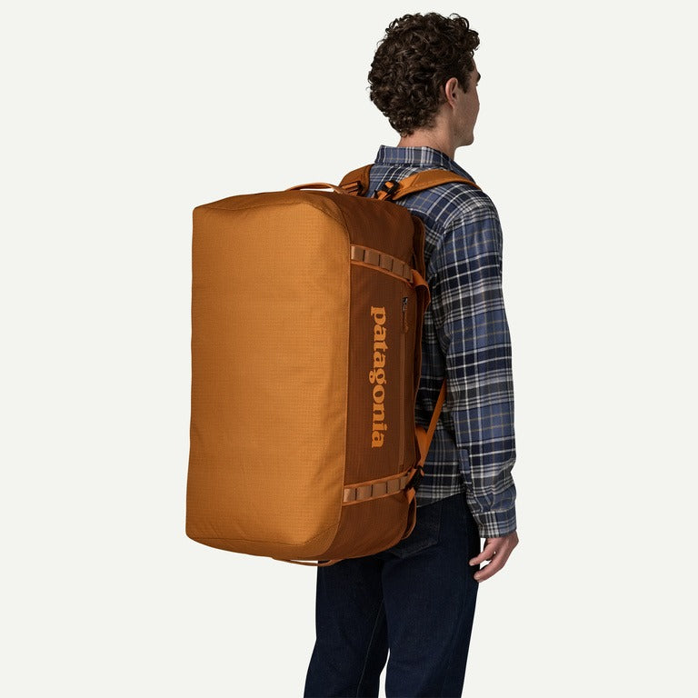 A man shown from the back wearing a large Patagonia duffel backpack in a warm camel brown color, paired with a blue plaid shirt and dark jeans, against a solid off-white background.