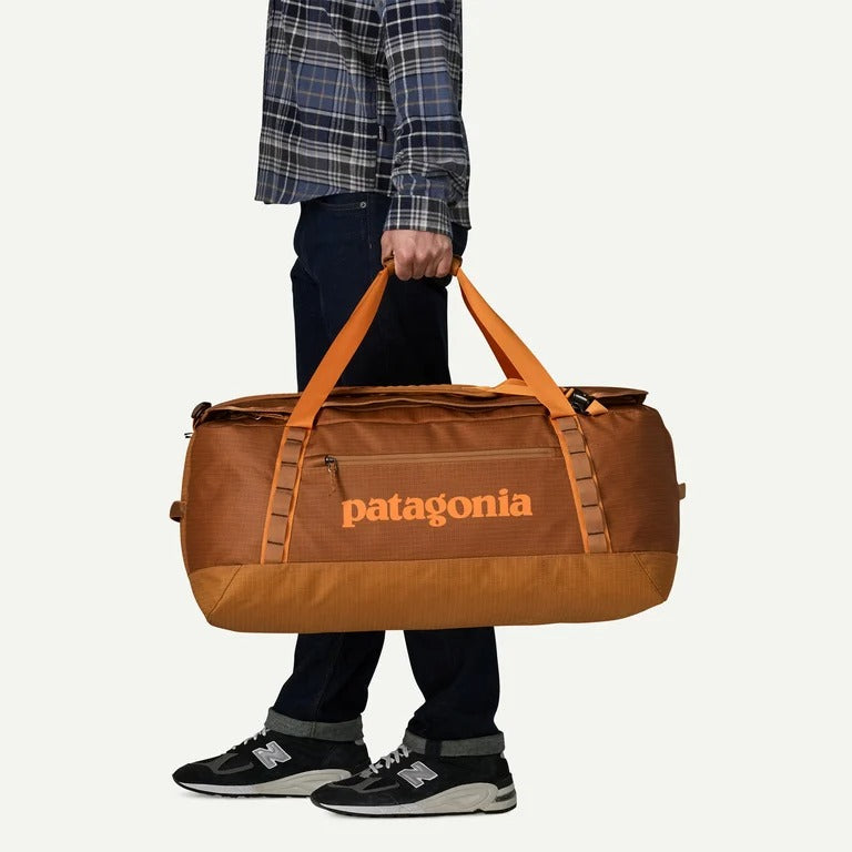 A person wearing a blue plaid shirt, dark jeans, and New Balance sneakers carries a large brown Patagonia duffel bag with orange handles and a bright orange Patagonia logo on the front, set against a plain off-white background.