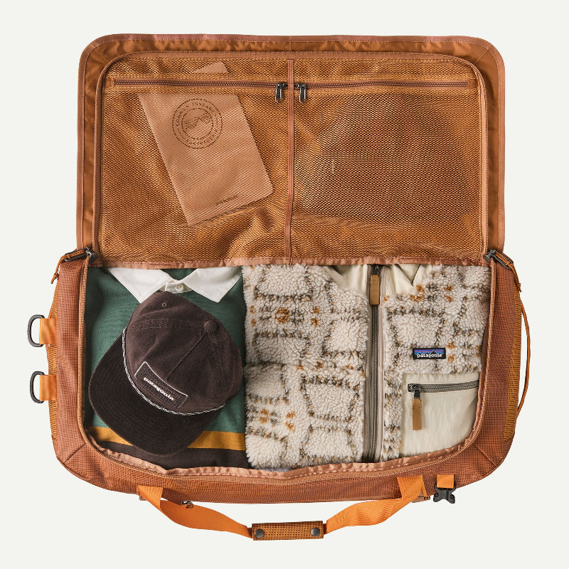 An open brown Patagonia suitcase or duffel bag shown from above, packed with clothing and accessories including a patterned fleece jacket, a dark cap, a green shirt, and a notebook. The Patagonia logo is visible on the jacket pocket and the cap.