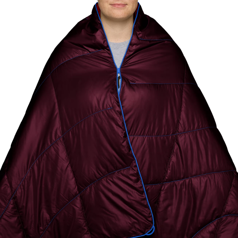 A person wrapped in a quilted puffer blanket or sleeping bag. The product is a deep burgundy/maroon color with bright blue piping and zipper trim, shown against a plain white background.