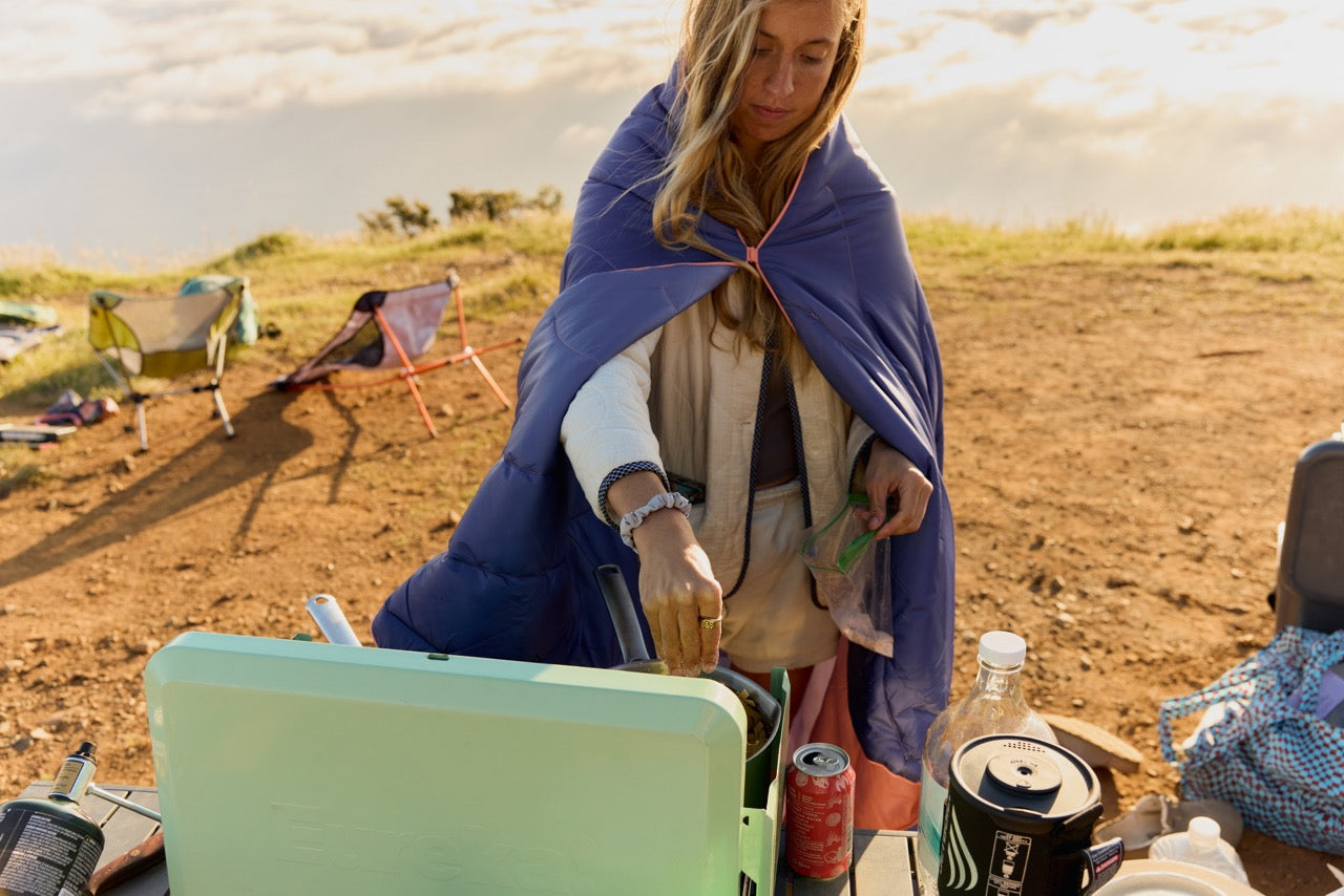 Person with a blue blanket over shoulders at a campsite with a cooler and drinks.
