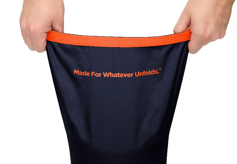 A navy blue bag with an orange waistband and orange text reading 'Made For Whatever Unfolds.' is being held open by two hands against a white background.