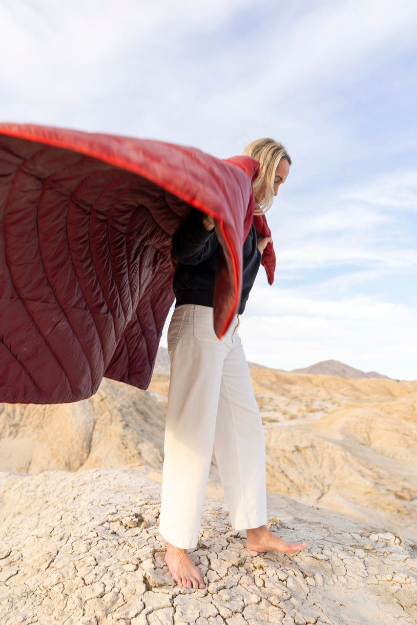 Woman in a red quilted rumpl waterproof blanket standing on rocky terrain with a mountainous background