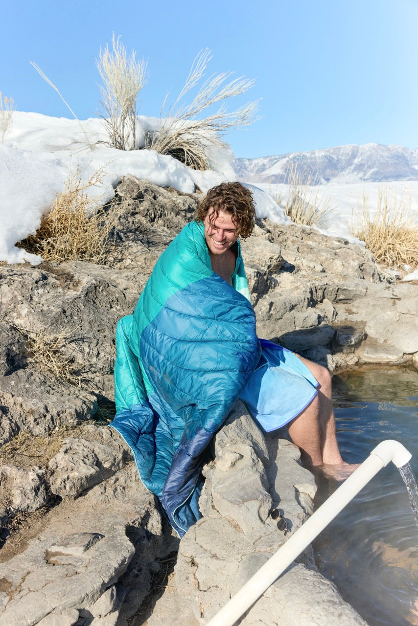 Male wrapped in a blue Rumply Travel Blanker next to a hot spring with mountains in the background
