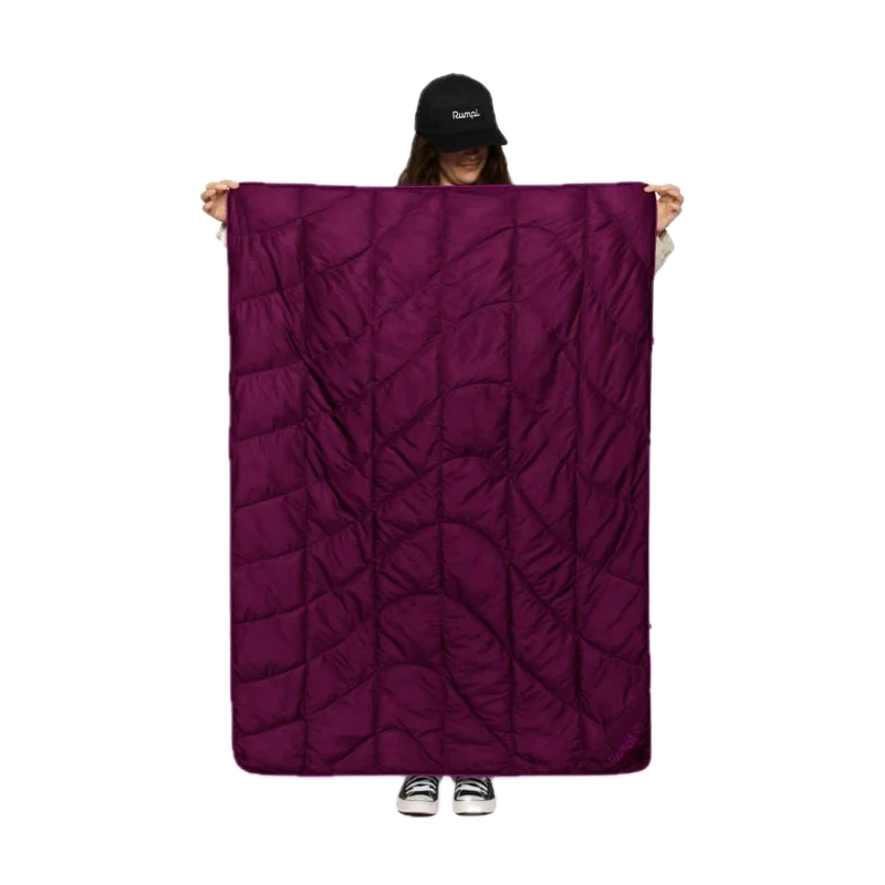A person wearing a black cap holds up a large, dark purple quilted sleeping pad or camping blanket against a solid white background.