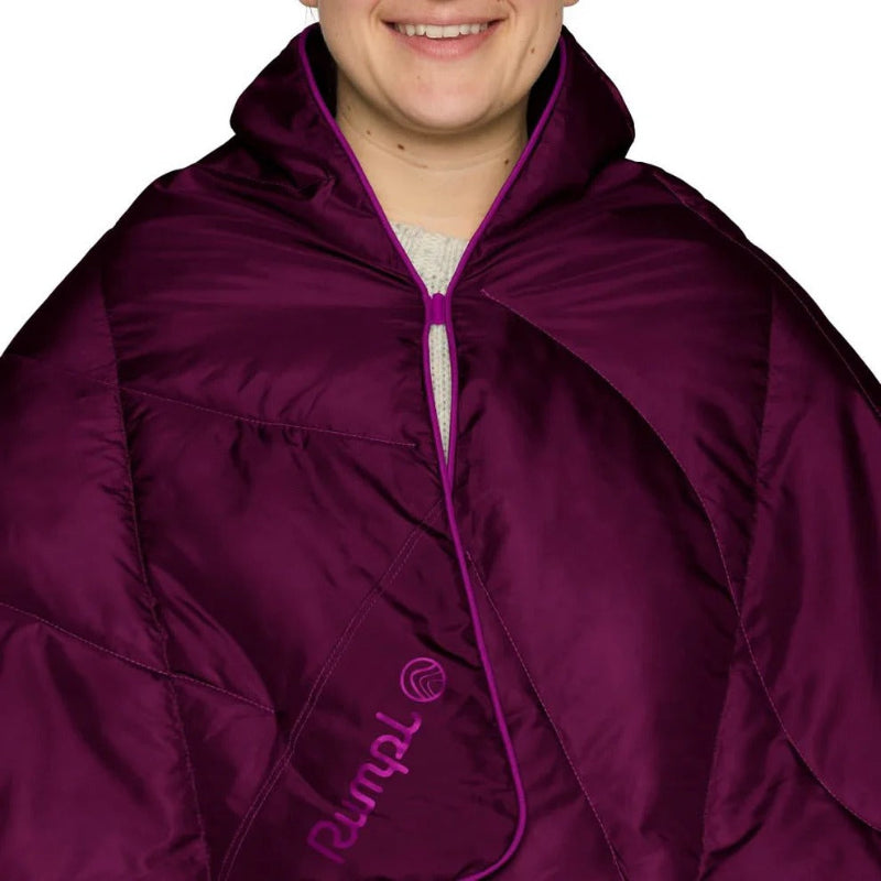 A close-up of a person wearing a deep purple puffer poncho or cape with a matching purple zipper and trim. The brand name 'Rumpl' is printed in purple on the lower left side.