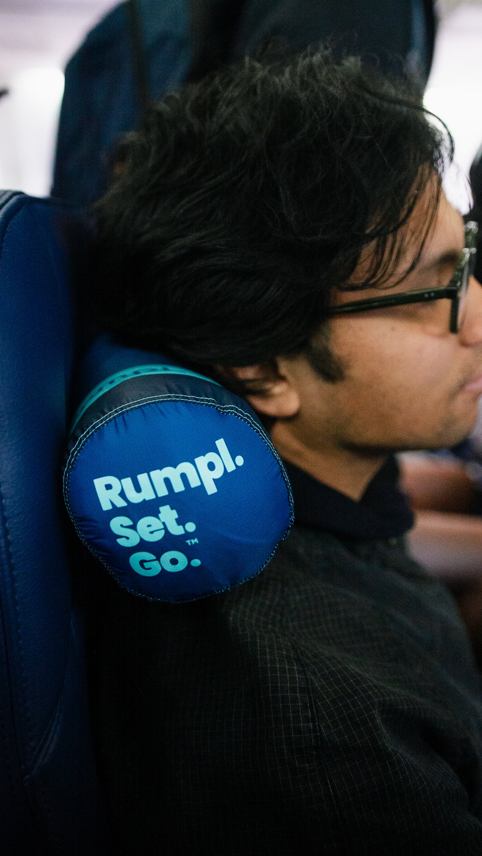 Person using a blue rumpl neck pillow with 'Rumpl. Set. Go.' text on an airplane seat
