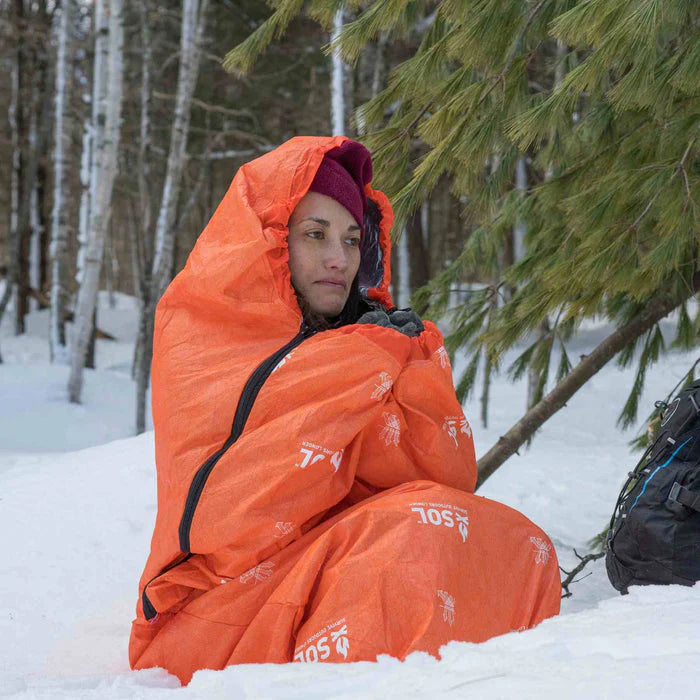 Person wrapped in an orange SOL Escape Bivvy in a snowy forest