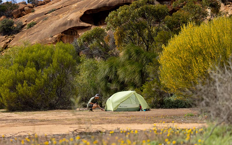 Person setting up a tent in a natural outdoor setting with trees and rocks.