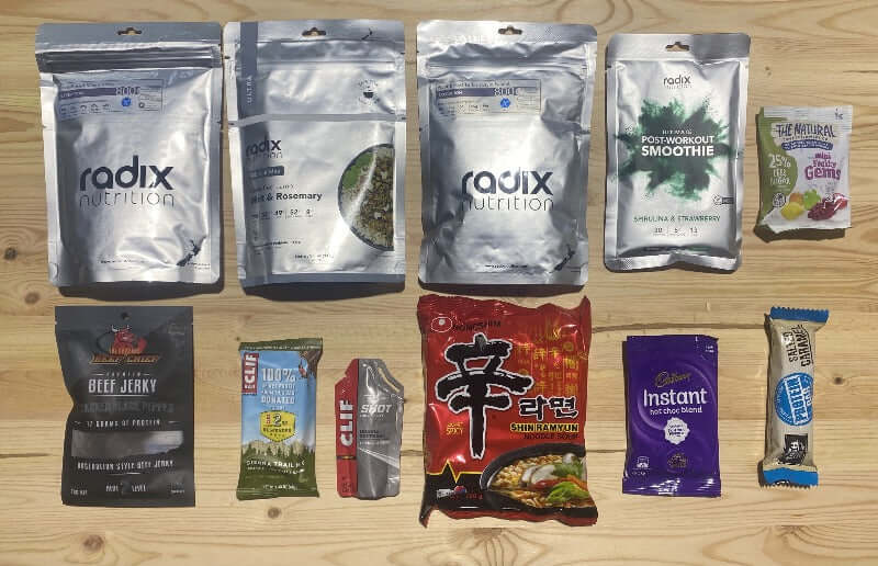 An array of packaged food items including meal packs and snacks spread out on a wooden surface, with brands such as Radix, Clif Bar, and The Original Beef Chief visible.